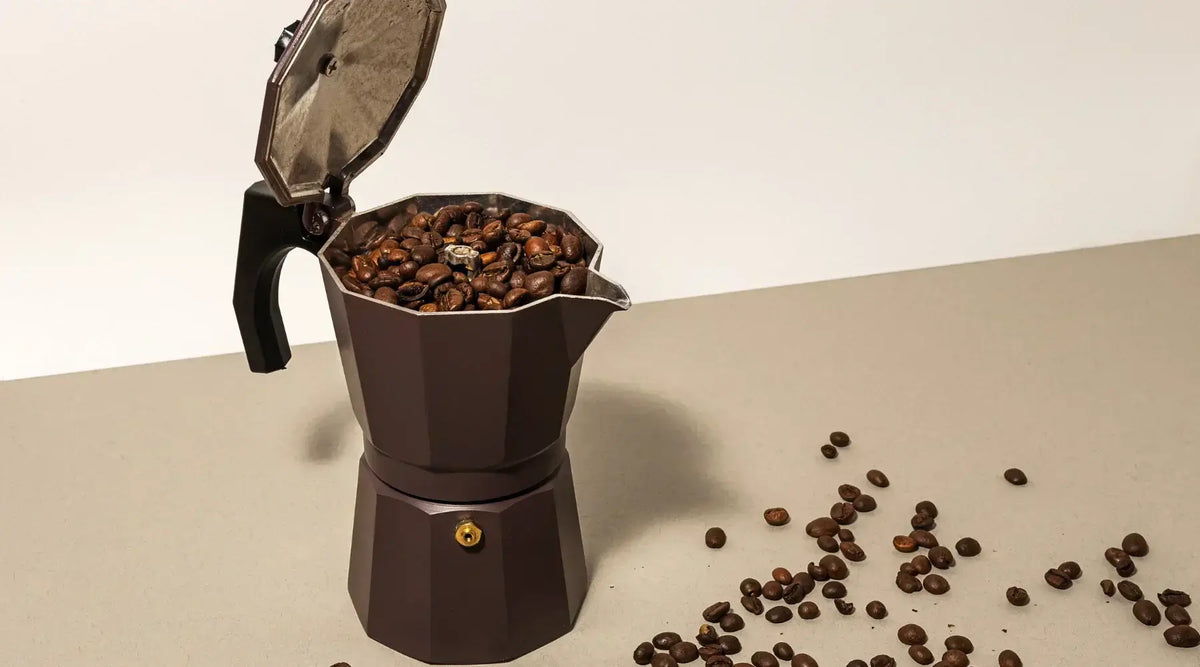 Cafetière italienne moka en aluminium posée sur un plan de travail, entourée de tasses de café fumant et de grains torréfiés.