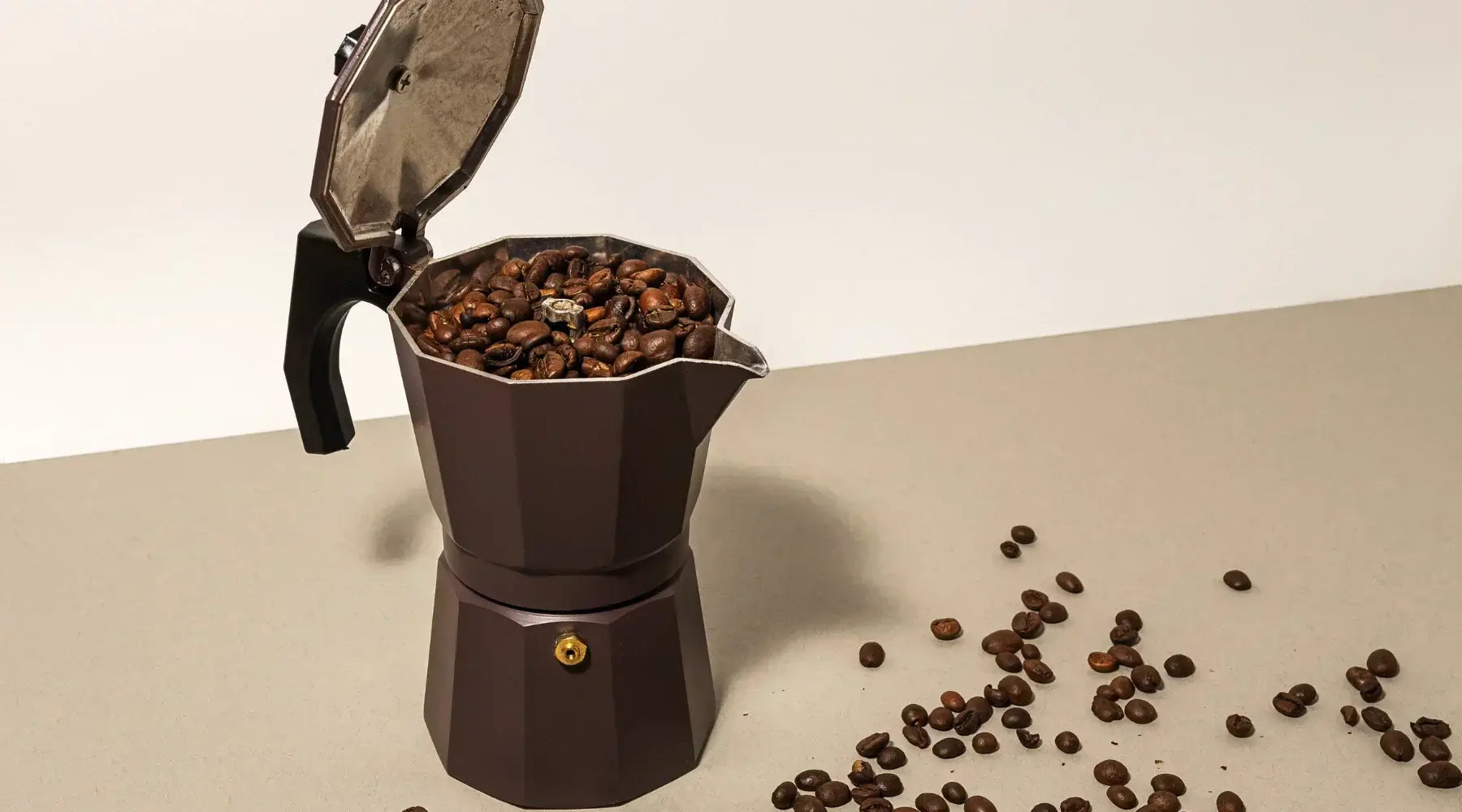 Cafetière italienne moka en aluminium posée sur un plan de travail, entourée de tasses de café fumant et de grains torréfiés.