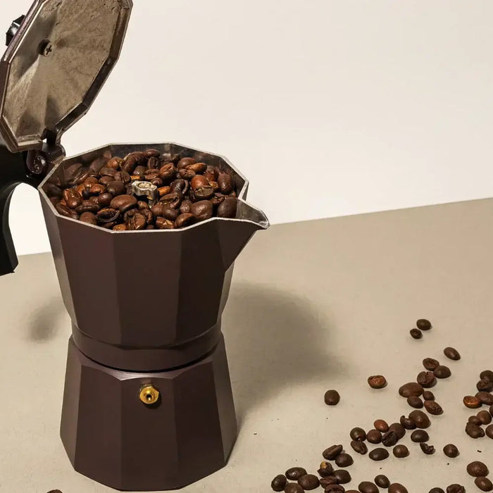 Cafetière italienne moka en aluminium posée sur un plan de travail, entourée de tasses de café fumant et de grains torréfiés.