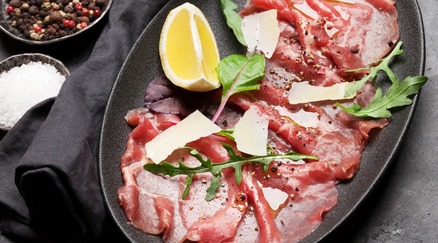 Carpaccio de bœuf servi sur une assiette noire, garni de roquette, copeaux de Grana Padano, citron et un filet d'huile d'olive, accompagné de grains de poivre et sel en arrière-plan.