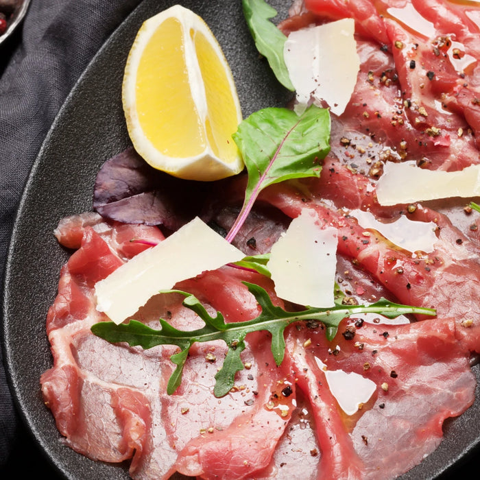 Carpaccio de bœuf servi sur une assiette noire, garni de roquette, copeaux de Grana Padano, citron et un filet d'huile d'olive, accompagné de grains de poivre et sel en arrière-plan.