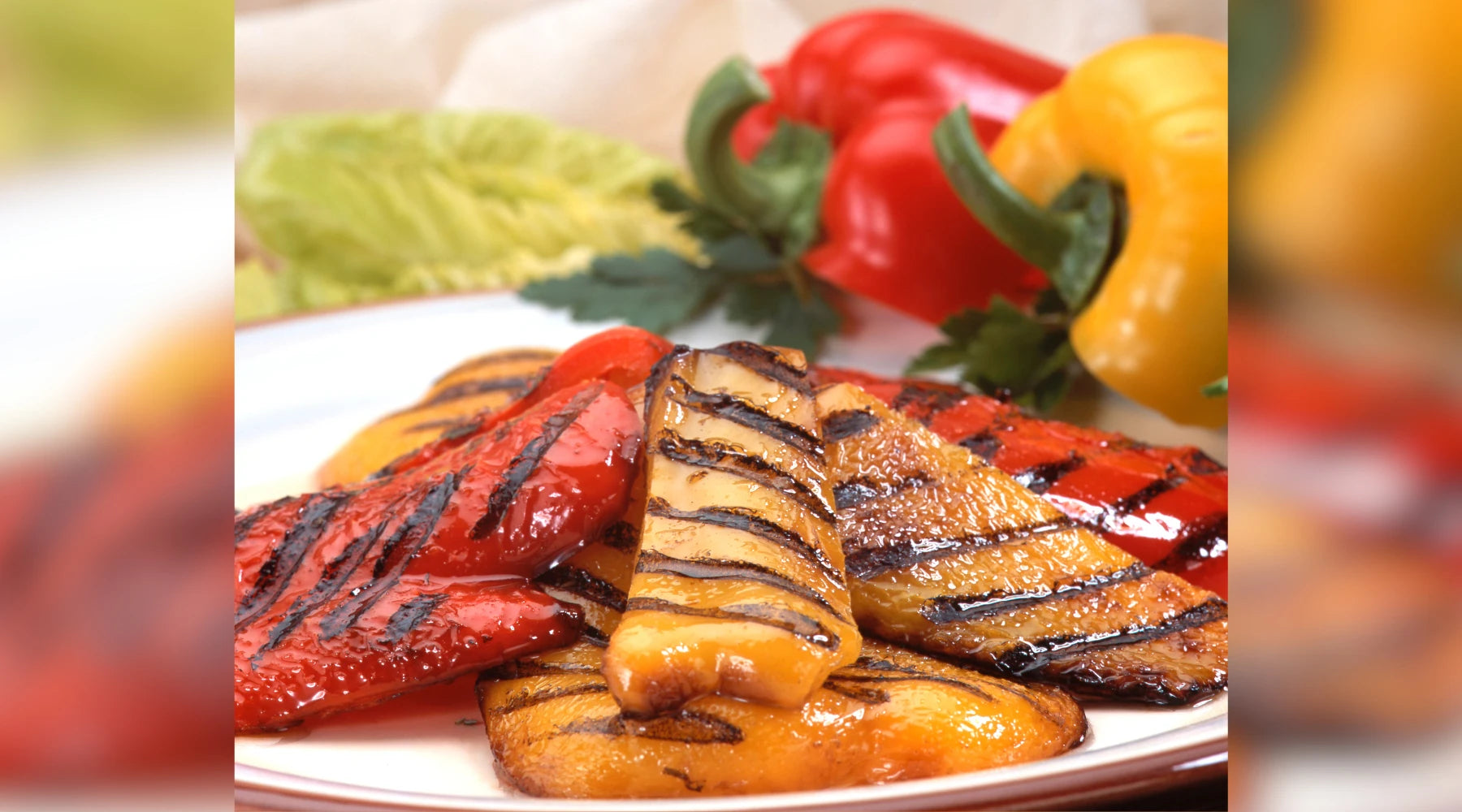 Assiette de poivrons grillés rouges et jaunes avec des marques de cuisson, accompagnée de feuilles de salade et de poivrons frais en arrière-plan.