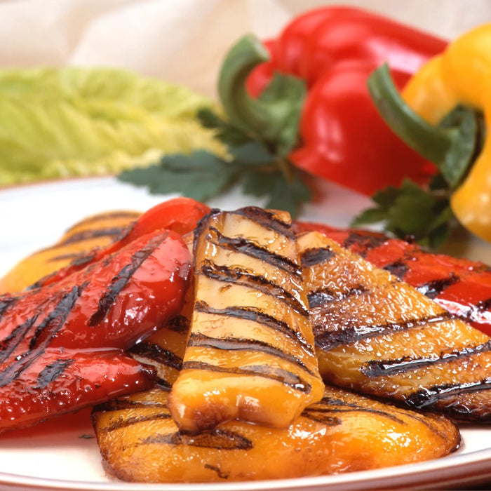 Assiette de poivrons grillés rouges et jaunes avec des marques de cuisson, accompagnée de feuilles de salade et de poivrons frais en arrière-plan.