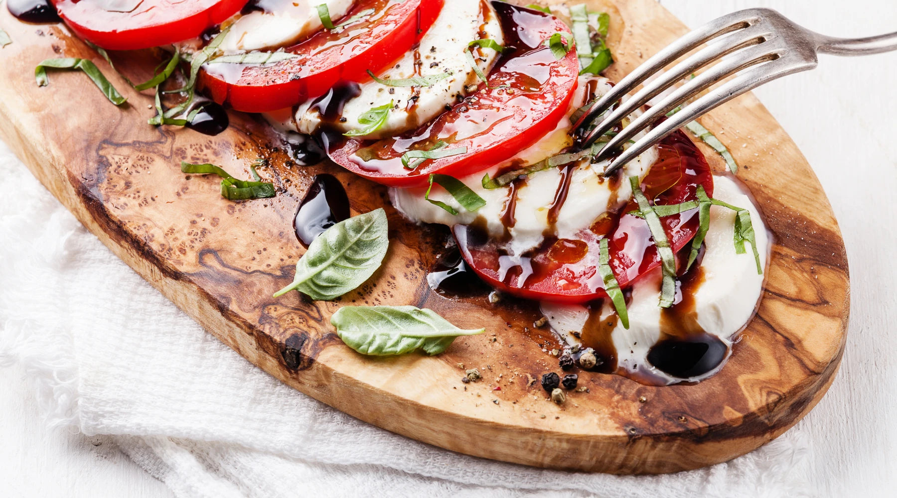 Salade Caprese sur une planche en bois, composée de rondelles de tomates et de mozzarella, garnie de basilic frais et arrosée de vinaigre balsamique, avec une fourchette en métal prête à servir.