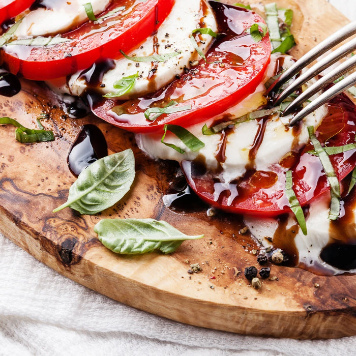 Salade Caprese sur une planche en bois, composée de rondelles de tomates et de mozzarella, garnie de basilic frais et arrosée de vinaigre balsamique, avec une fourchette en métal prête à servir.