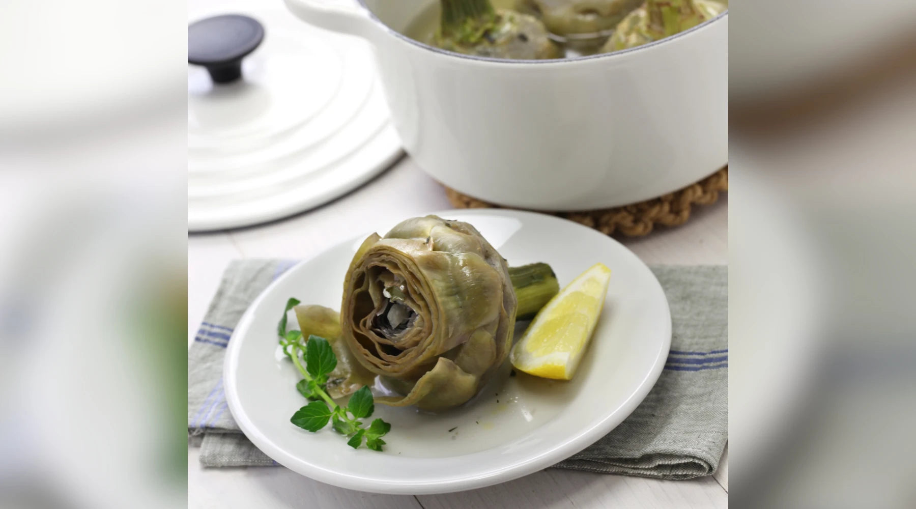 Artichaut à la romaine servi sur une assiette blanche avec une tranche de citron et des herbes fraîches, accompagné d'une cocotte en arrière-plan.