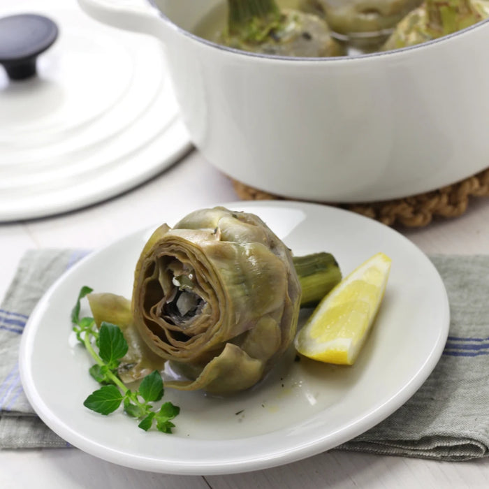 Artichaut à la romaine servi sur une assiette blanche avec une tranche de citron et des herbes fraîches, accompagné d'une cocotte en arrière-plan.